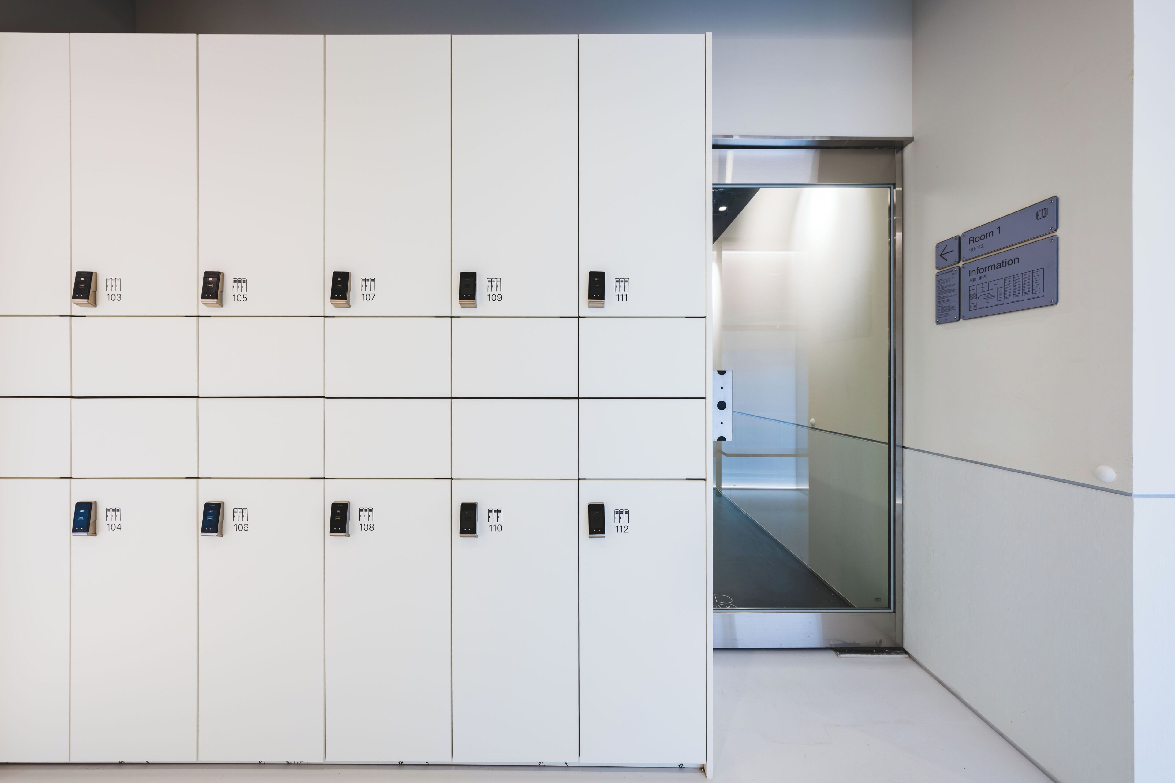 Une rangée de casiers blancs numérotés de 103 à 112, un ascenseur en verre et un panneau indiquant « Bureau d’information, salle 1 ».