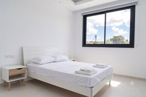 A white bedroom with a bed, a nightstand, a window with a view, and a ceiling light. - Sykia | Upper Floor 2-BR Apartment | (Paphos)