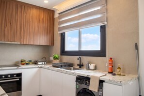 A modern kitchen with wooden cabinets, a built-in oven, a dishwasher, a window with blinds, and a countertop with various items. - Sykia | Upper Floor 2-BR Apartment | (Paphos)