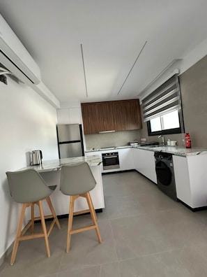 A modern kitchen with white cabinetry, a built-in oven, a dishwasher, and a coffee machine. There are two bar stools and a window with blinds. - Sykia | Upper Floor 2-BR Apartment | (Paphos)