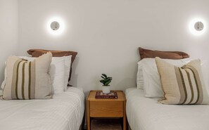 Ein Schlafzimmer mit zwei Betten, jeweils mit Kopfkissen und Decke, einem Nachttisch mit Buch und Pflanze sowie zwei Wandleuchten.