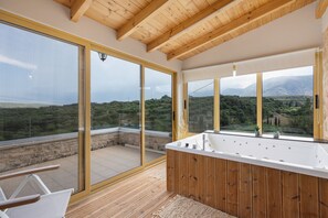 A modern bathroom with a large bathtub, wooden flooring, and large windows offering a view of the countryside. - Cretan Traditional Home Villa with Mountain View, Wi-Fi, and Air Conditioning (Tilisos)