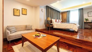 A hotel room with a sofa, a wooden coffee table with fruit, a bed with a white comforter, and a large window with curtains. - Luxurious retreat with premium amenities & comfort (Bangkok)