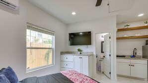 Un dormitorio con una cama, un televisor montado en la pared, una ventana y una zona de cocina con armarios y una encimera.