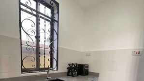 A kitchen with a sink, cabinets, a window with decorative bars, and a tiled floor. - Cozy Studio Near City & Masaki (Dar es Salaam)