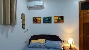 A bedroom with a bed, a nightstand, a lamp, an air conditioner, and three wall-mounted pictures. - Cozy Studio Near City & Masaki (Dar es Salaam)