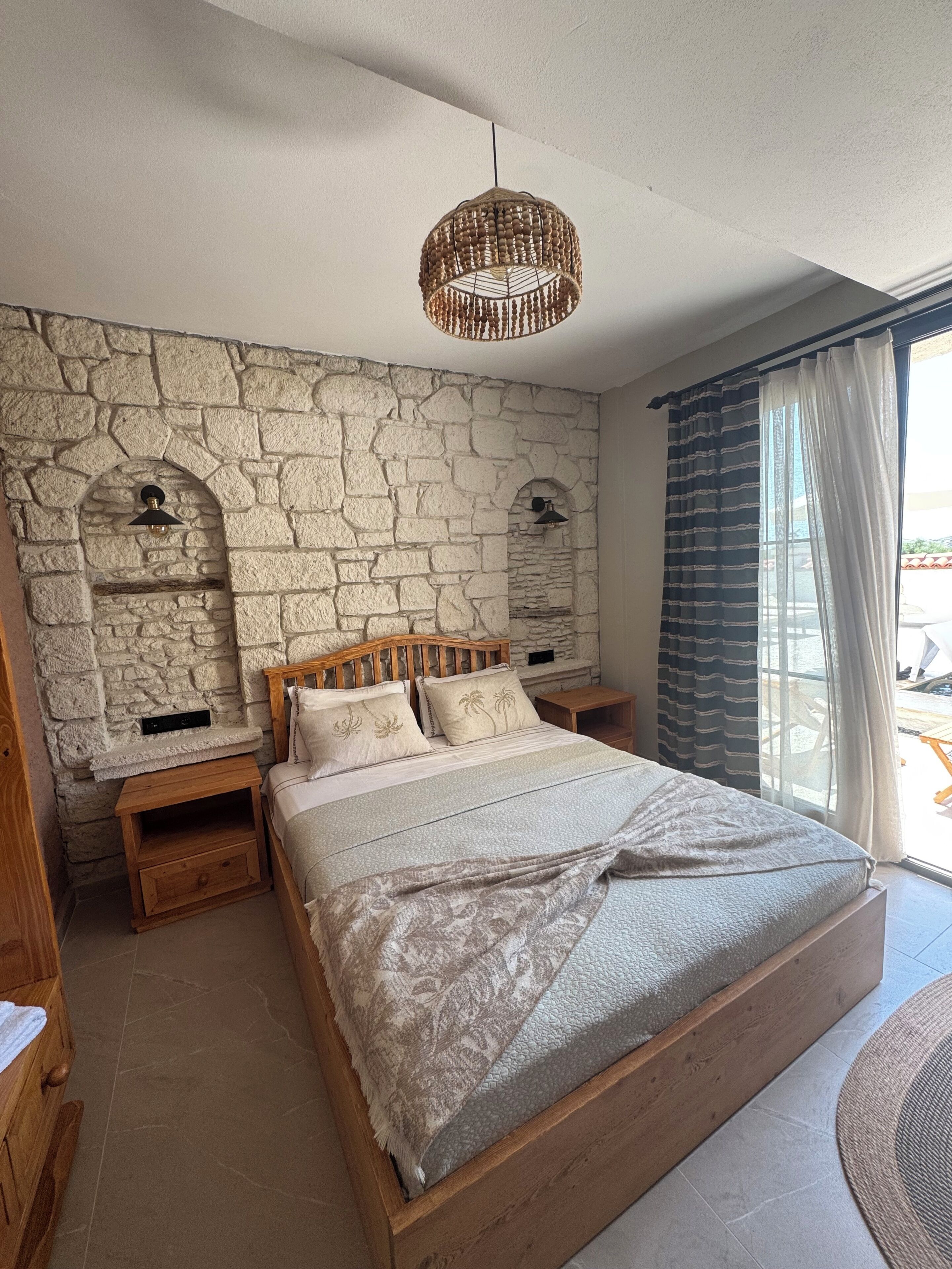Une chambre avec un mur en pierre, un lit recouvert d’une couvre-lit à motifs, une tête de lit en bois, des tables de chevet et un luminaire suspendu.