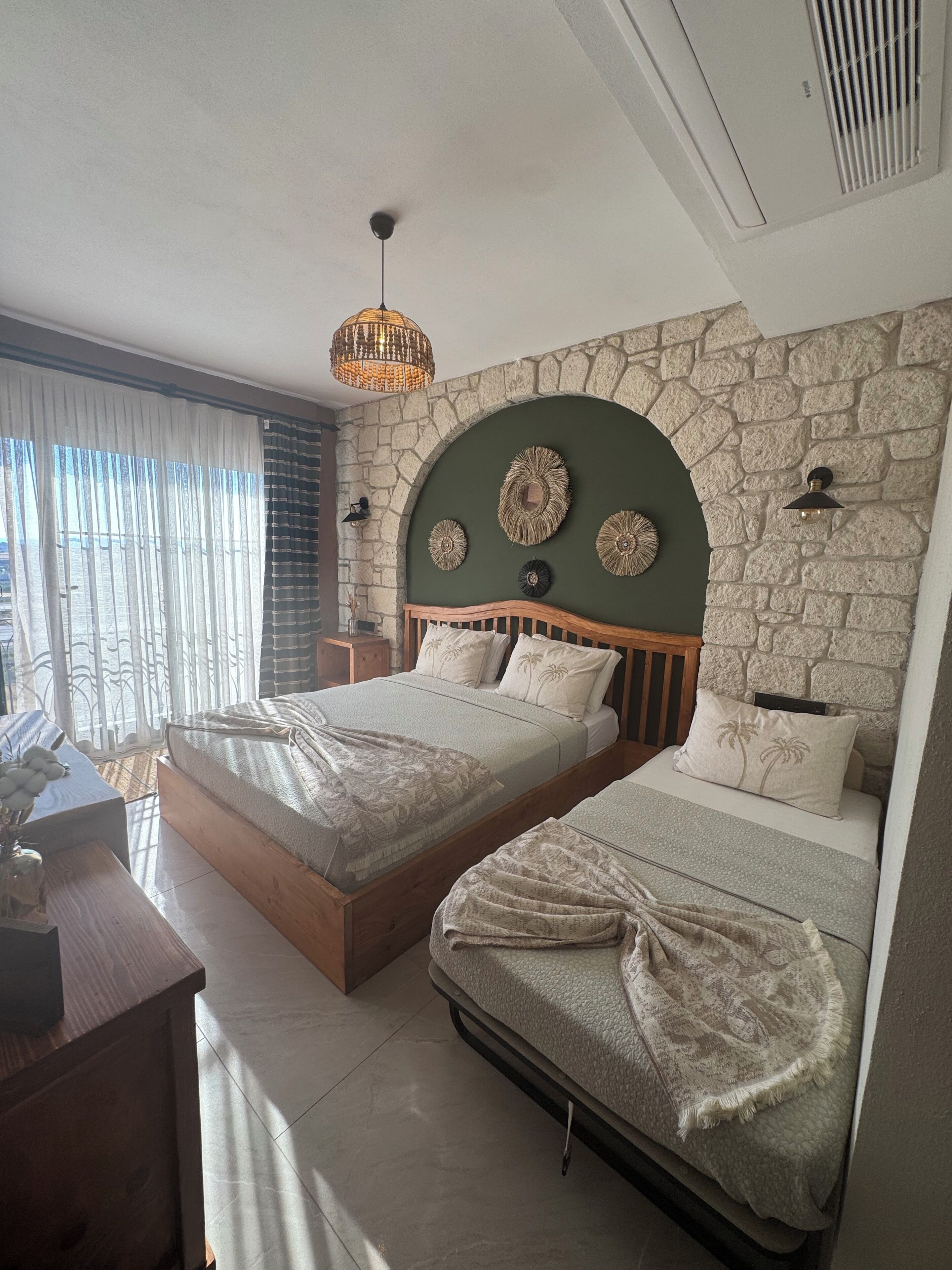 Une chambre avec deux lits, un mur en pierre, un luminaire suspendu et une vue par la fenêtre.