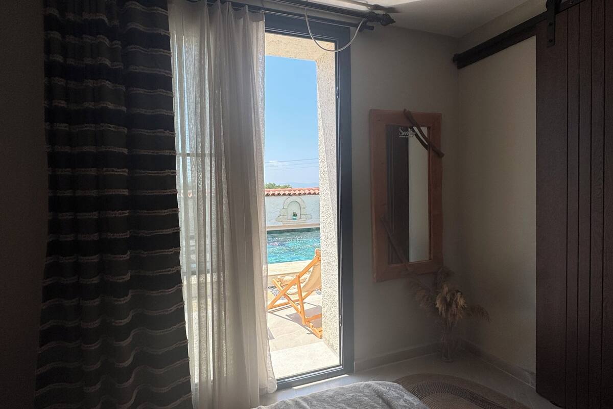 A bedroom with a bed, a window with curtains, a view of the outside, and a door leading to a balcony.