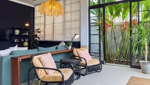 Un salon moderne comprenant un canapé, deux fauteuils, un ventilateur de plafond, un luminaire suspendu et de grandes fenêtres ornées de plantes tropicales.