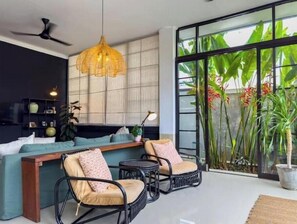 A modern living room with a sofa, two chairs, a ceiling fan, a hanging light fixture, and large windows with tropical plants. - Rumah Rama 3BR Private Pool Villa• Surrounded by Best Beaches in Bali (Kuta utara)