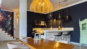 Une cuisine moderne avec un coin repas, comprenant une table en bois, des chaises, des luminaires suspendus et un escalier orné d’une fresque florale.