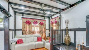 Uma sala de estar com um sofá branco, almofadas vermelhas, piso de madeira, tapete vermelho, ventilador de teto e escada decorada com um vaso e velas.