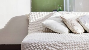 Un sofá blanco con textura y un cojín, una pared verde y una ventana con una planta.