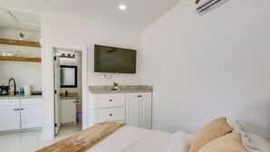 Un dormitorio con cama, televisor montado en la pared, baño con lavamanos y espejo, y una zona de cocina con armarios y encimera.