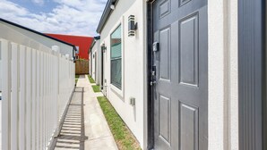 Un étroit passage entre deux bâtiments, avec une clôture blanche d’un côté et une porte grise de l’autre.