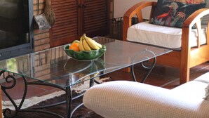 Een glazen tafel met een schaal fruit, een houten stoel met kussen en een rieten mand op een betegelde vloer.
