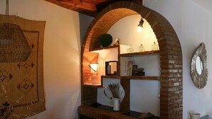 Een gezellige kamer met een bakstenen boog, een geweven wanddecoratie, een plank met boeken en flessen, en een decoratieve spiegel.