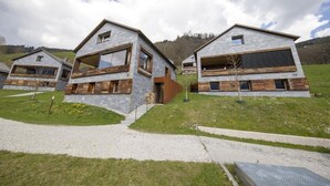 Une maison moderne à deux étages, dotée d’une façade en pierre et de détails en bois, située dans un espace verdoyant sous un ciel dégagé.