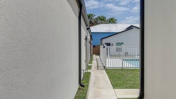Un étroit sentier mène à un espace piscine, délimité par une clôture blanche, et à un bâtiment surmonté d’un toit bleu.