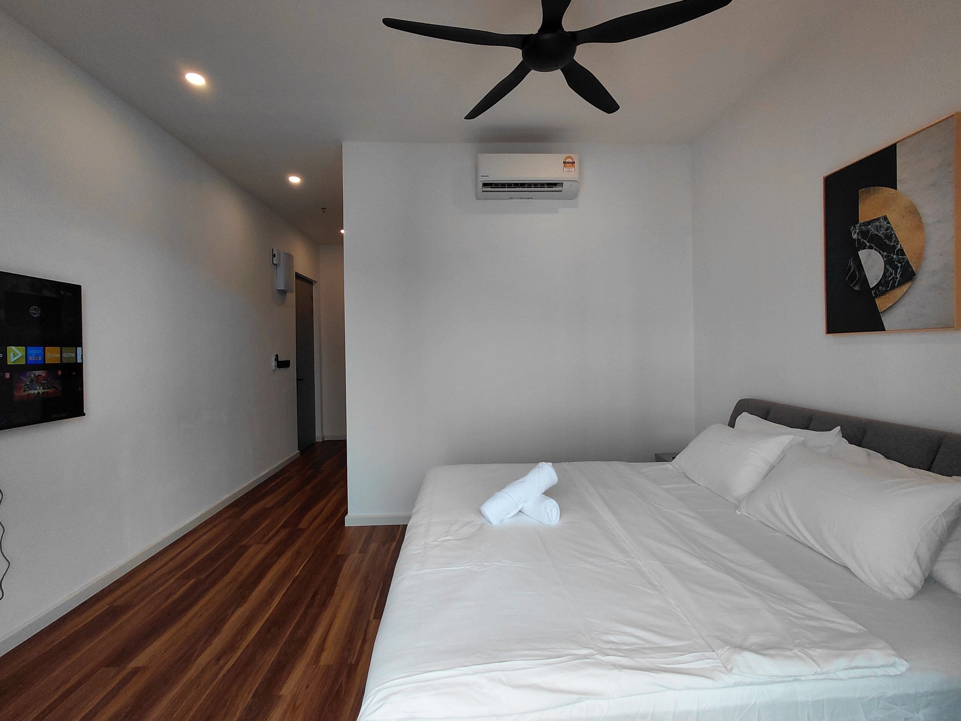 Habitación de hotel con cama, ventilador de techo, equipo de aire acondicionado, televisor de pantalla plana y pasillo con piso de madera.
