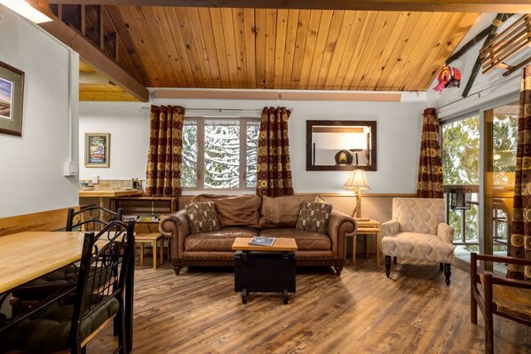 A cozy living room with a wooden ceiling, a brown leather sofa, a small table, and a chair. - Rustic Charm Condo #16 at Ptarmigan Village (Whitefish)