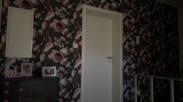A room with a floral wallpaper, a closed door, a dresser with a 'Welcome' sign, and a picture frame.