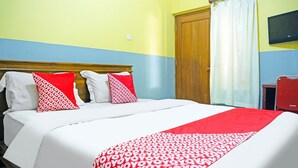 Habitación de hotel con una cama, dos almohadas, una colcha roja y blanca, cabecera de madera, puerta de madera, televisor en la pared y una ventana con cortinas.