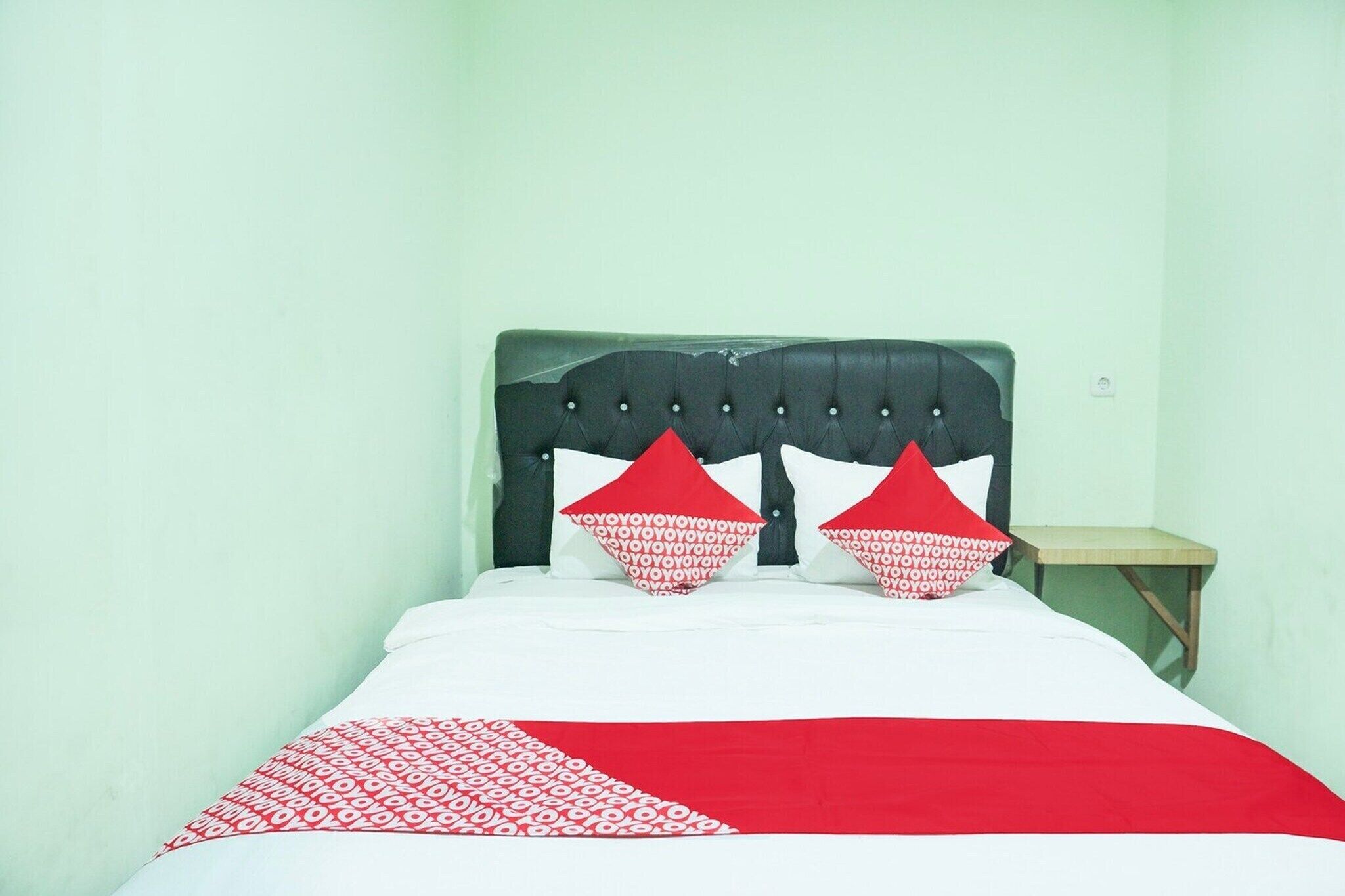 Een bed met een zwart hoofdeinde, wit-rode beddengoed en een klein houten nachtkastje ernaast.