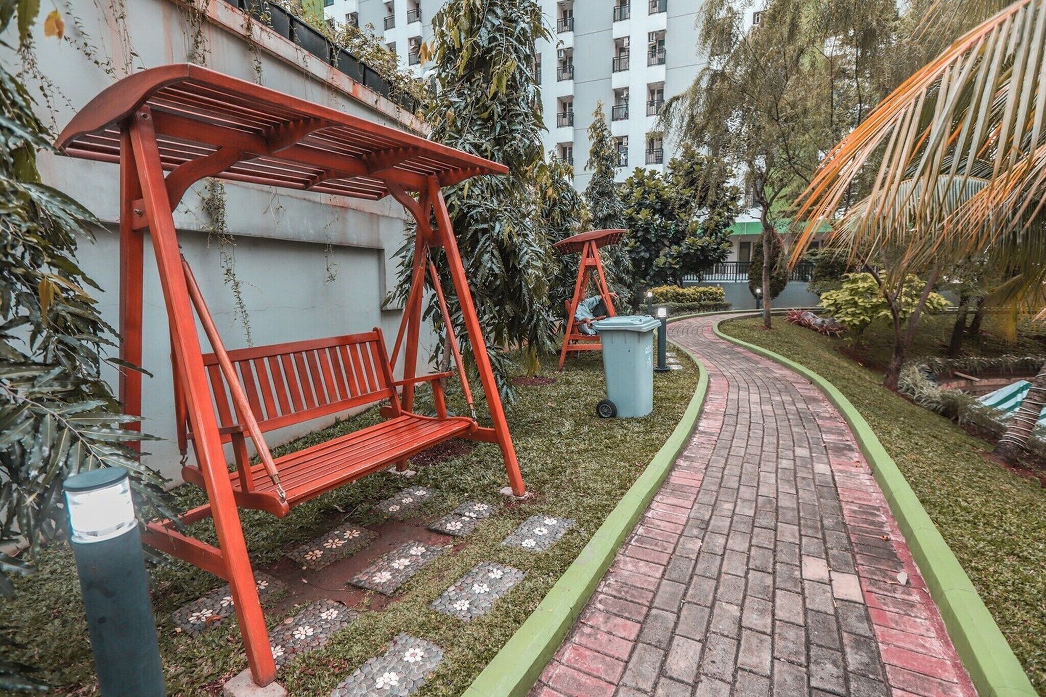 A pathway leading to a red bench with a metal frame, surrounded by greenery and a brick path.