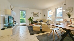 Een moderne woonkamer met een bank, salontafel en eettafel, een televisie en een groot raam.