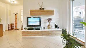 Een moderne woonkamer met een flatscreen televisie, een houten console tafel, een vaas met gedroogde bloemen en een groot raam met uitzicht op buiten.