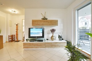 Een moderne woonkamer met een flatscreen televisie, een houten console tafel, een vaas met gedroogde bloemen en een groot raam met uitzicht op buiten.