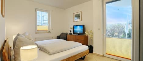 A bedroom with a bed, a TV, a window, a radiator, and a balcony.