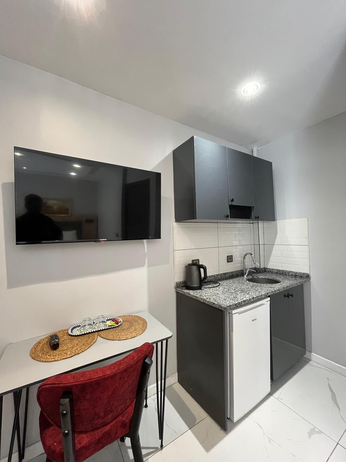 A modern kitchen with a flat-screen TV, a small dining table with a chair, and a coffee maker.