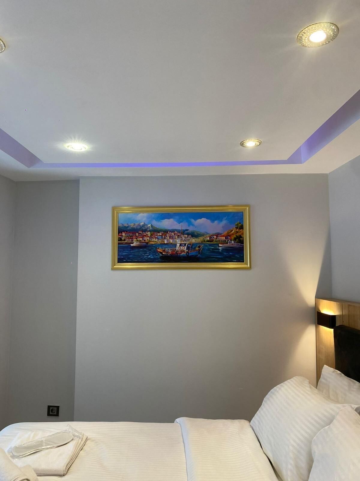 A hotel room with a bed, a painting of a coastal town, recessed ceiling lights, and a wall-mounted lamp.