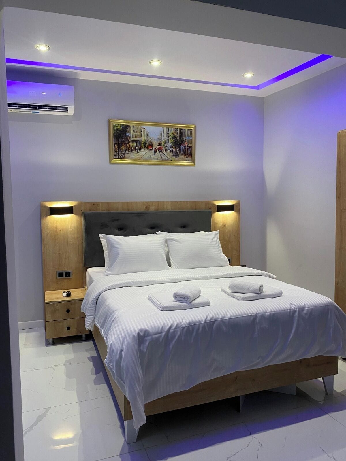 A modern bedroom with a wooden headboard, white bedding, a painting above the bed, and a nightstand with a phone and charger.