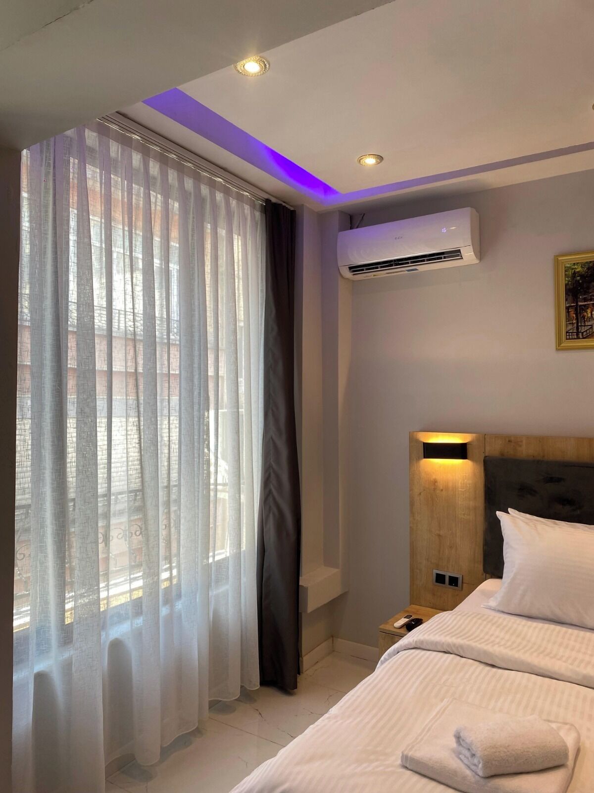 A hotel room with a bed, an air conditioning unit, a nightstand, and a window with sheer curtains.
