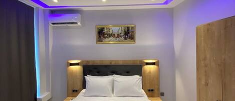 A hotel room with a bed, nightstands, a headboard, a painting, an air conditioning unit, and a wardrobe.