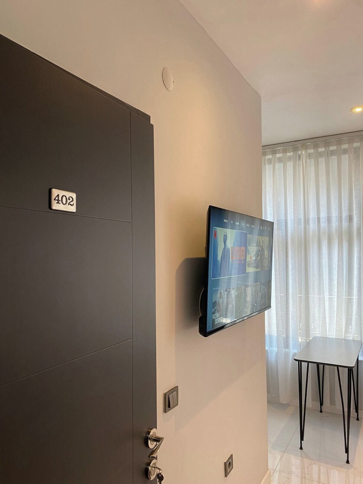 Habitación de hotel con un televisor de pantalla plana grande montado en la pared, una puerta con placa de número y una pequeña mesa con sillas.