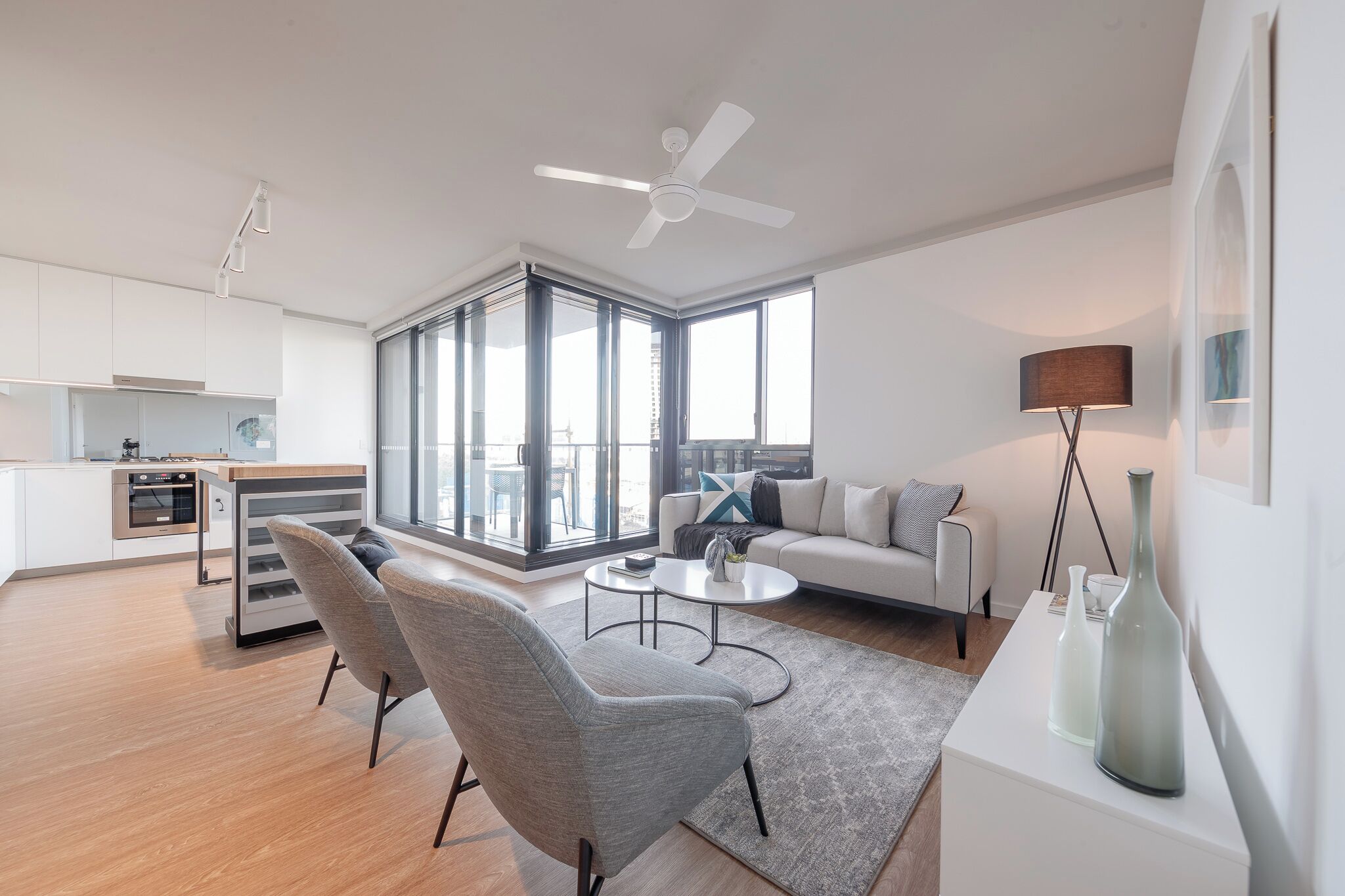 Una sala de estar moderna con sofá, mesa de centro y sillas, una cocina equipada y ventanas amplias con vistas.