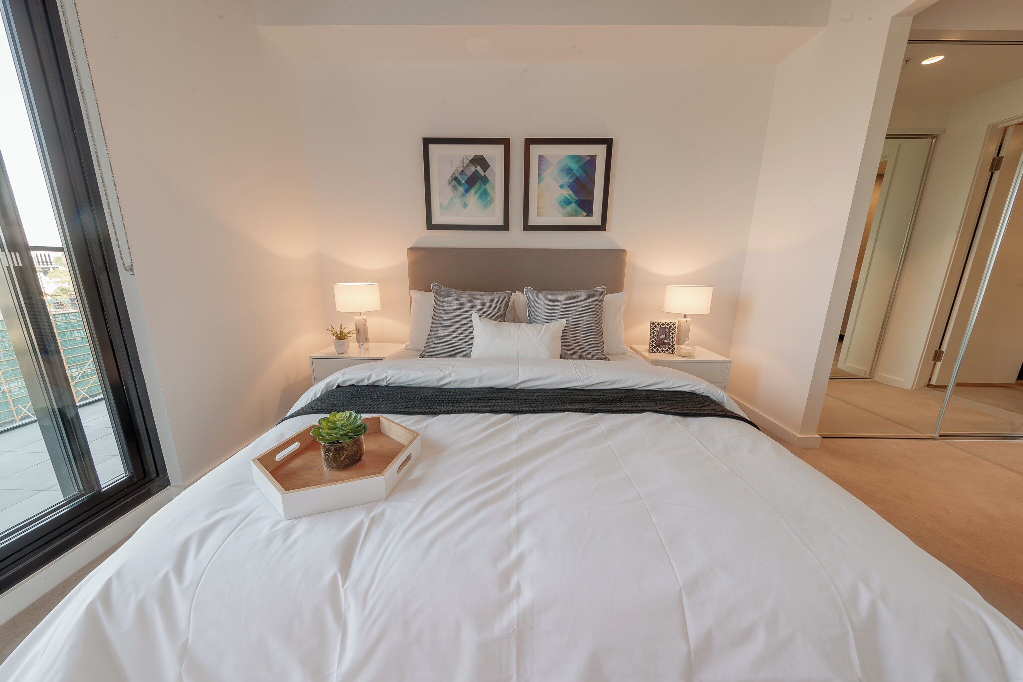 A modern hotel room with a large bed, two bedside tables with lamps, a window with a view, and two framed artworks on the wall.