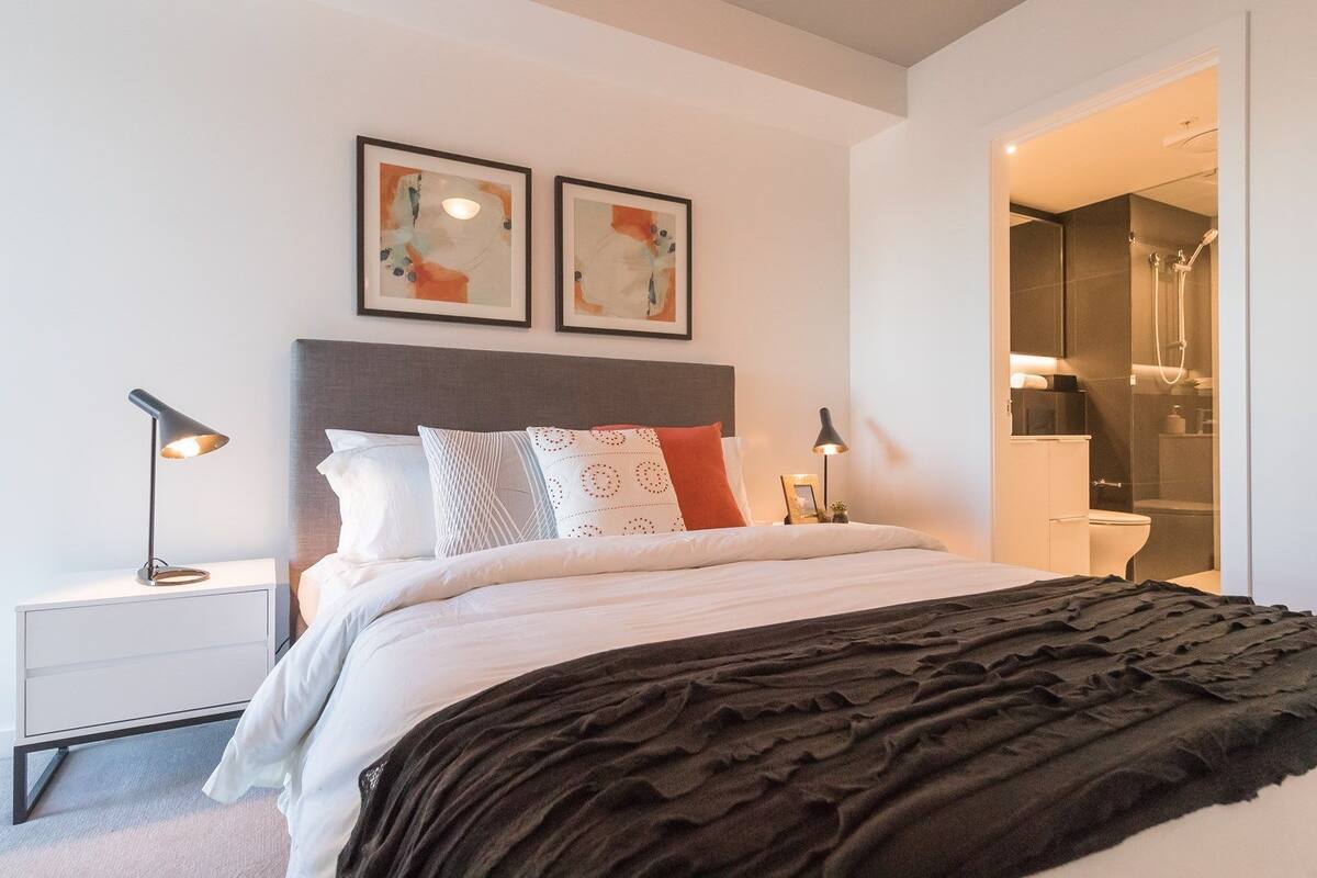 Un dormitorio moderno con una cama, mesitas de noche, un cabecero, dos obras de arte enmarcadas, y un baño visible a través de una puerta abierta.