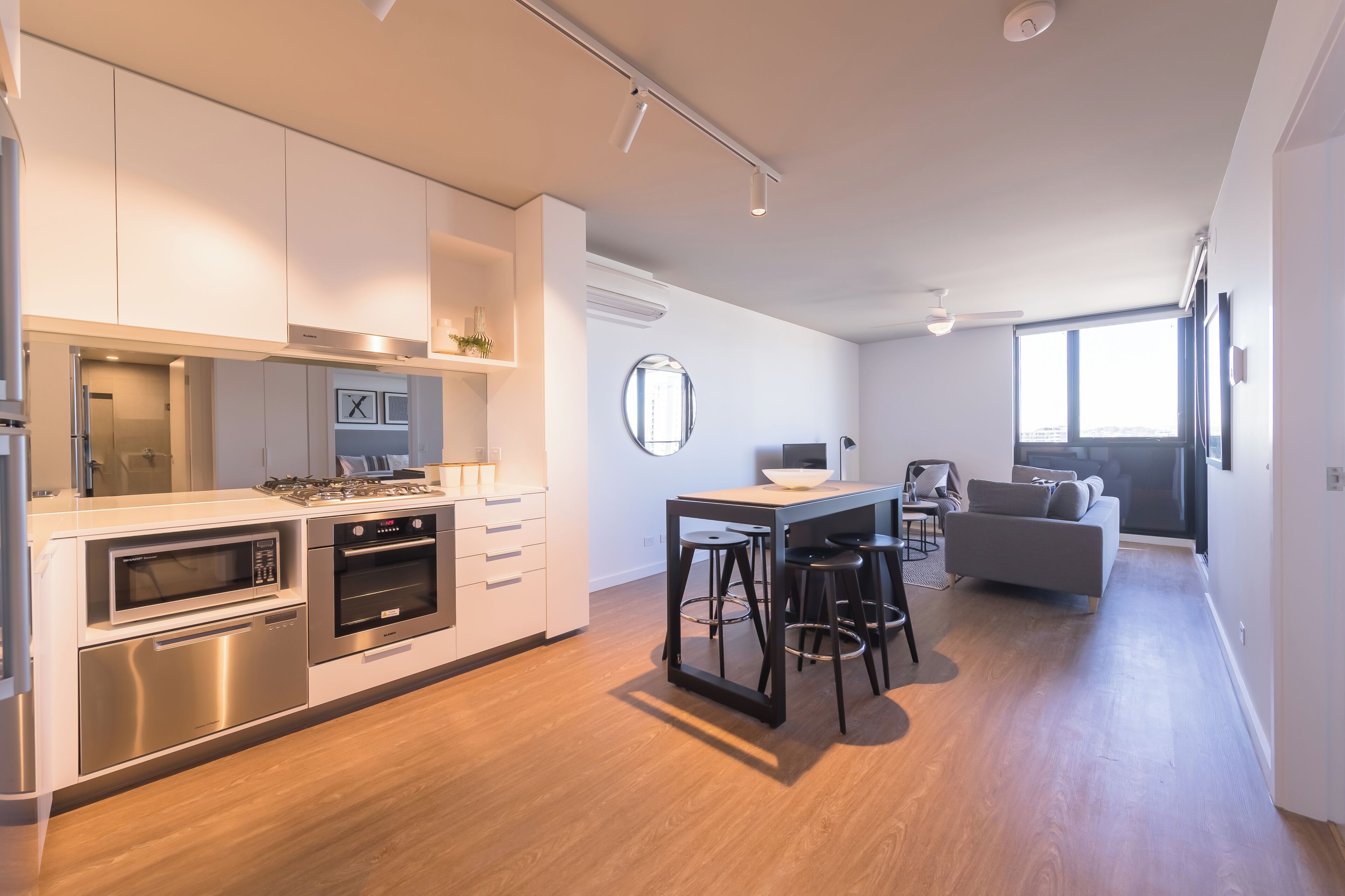 A modern kitchen with built-in appliances, a dining area with stools, and a grey sofa in a bright living space with large windows.