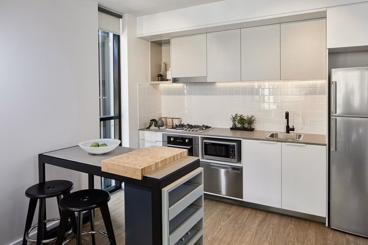 A modern kitchen with white cabinets, a stainless steel refrigerator, a built-in oven, and a small island with stools.