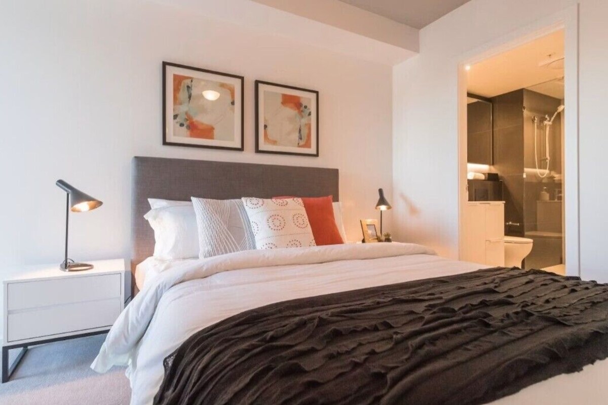 Un dormitorio moderno con una cama, lámparas de noche, una mesita de noche y un baño visible a través de una puerta abierta.