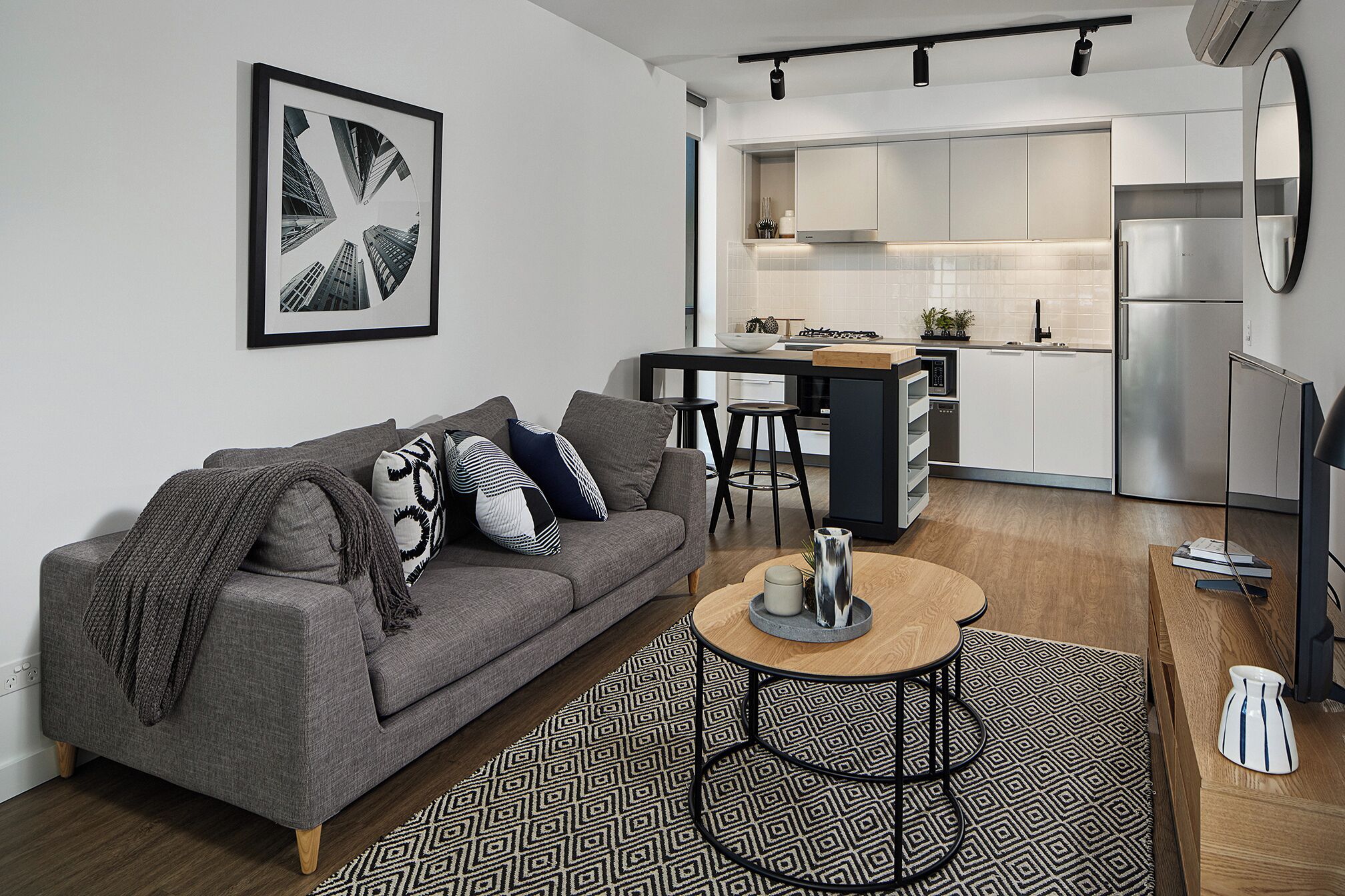 A modern living room with a grey sofa, a coffee table, and a kitchen area with white cabinets and a stainless steel refrigerator.