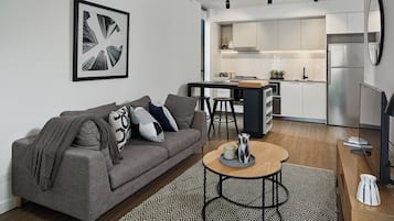 A modern living room with a grey sofa, a coffee table, and a kitchen area with white cabinets and a stainless steel refrigerator.
