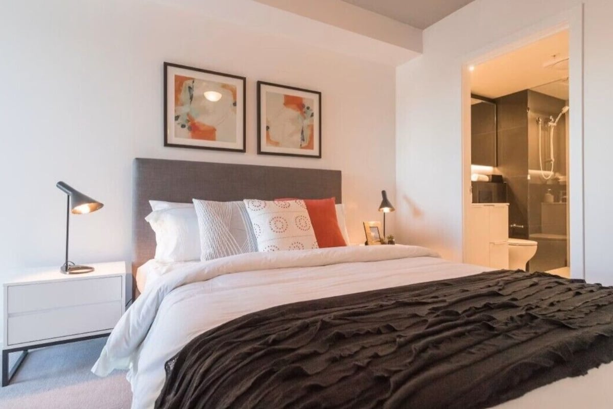 Un dormitorio moderno con una cama, lámparas de noche, una mesita de noche y un baño visible a través de una puerta abierta.