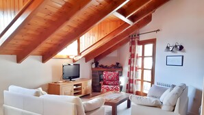 Ein Loft-Wohnzimmer mit Holzbalken, einem Fernseher, zwei weißen Sofas, einem Kamin und einem Fenster mit Vorhängen.